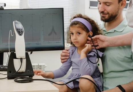 Criança realizando um exame de audiometria com a ajuda de um profissional e do pai.